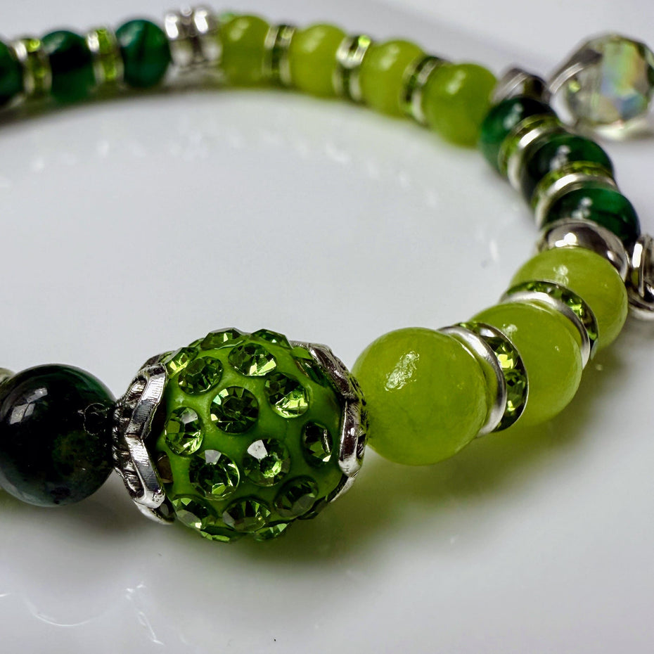 Frühlingshaftes Damenarmband mit strahlend grünen Perlen, glitzernden Strass-Elementen und filigranem Feder-Anhänger. Handgefertigt in der Schweiz, ideal für leichte Frühlingsoutfits.