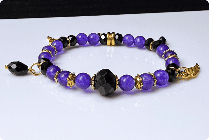 Elegantes Armband ‚Lila Traum‘ mit leuchtend lila Steinperlen, facettierten schwarzen Glasperlen und goldenen Akzenten. Verziert mit Anhängern wie einem goldenen Mond und einem Engelsflügel – ein luxuriöses, handgefertigtes Schmuckstück aus der Schweiz.