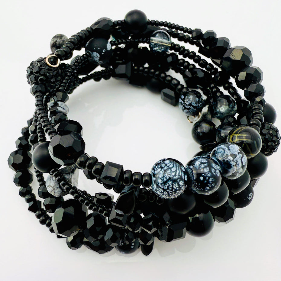 Elegantes spiralförmiges Armband mit einer vielseitigen Kombination aus schwarzen Glasperlen in verschiedenen Größen und Oberflächen. Handgefertigt in der Schweiz, flexibel ohne Verschluss, perfekt für stilvolle Alltagslooks und besondere Anlässe.