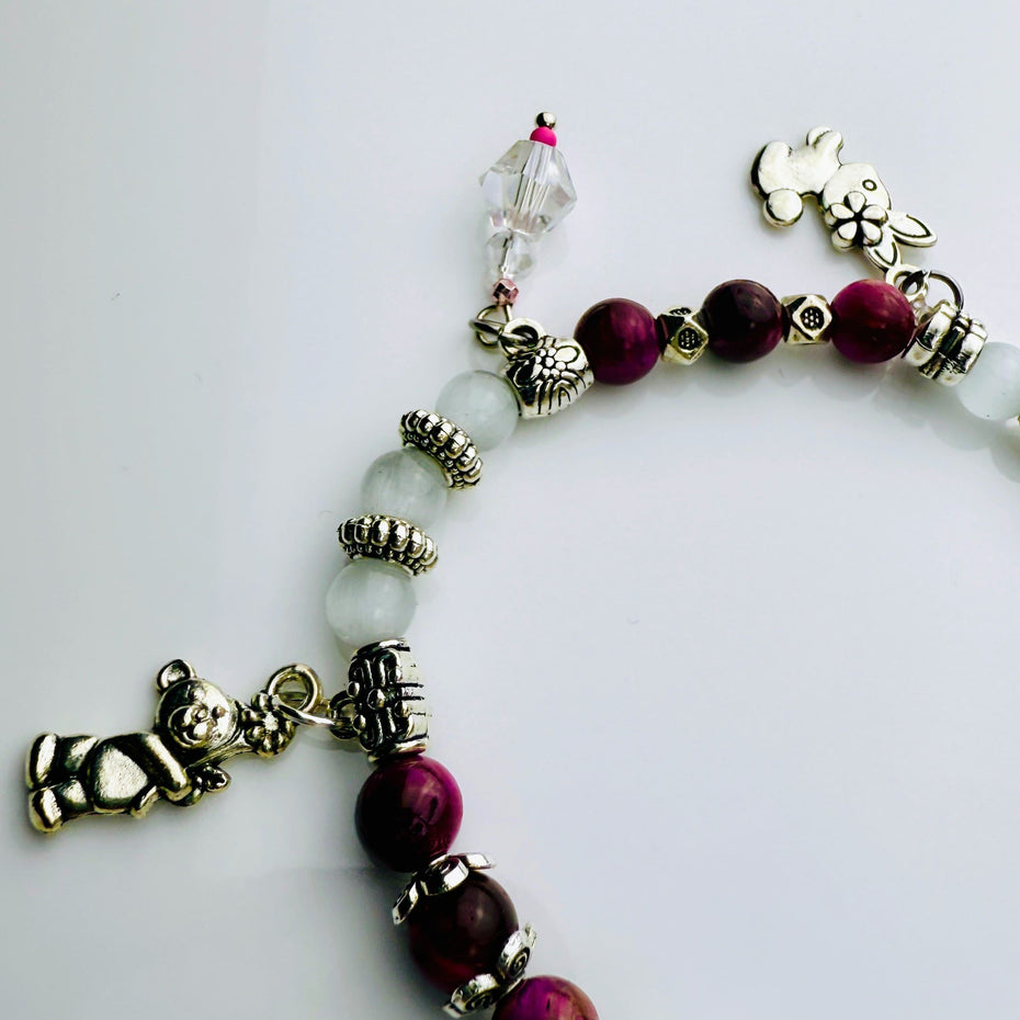 Verspieltes Damenarmband mit Anhängern wie Teddybär, Blüten und Schmuckelementen, kombiniert mit weißen und weinroten Perlen, ideal als verspieltes Accessoire.