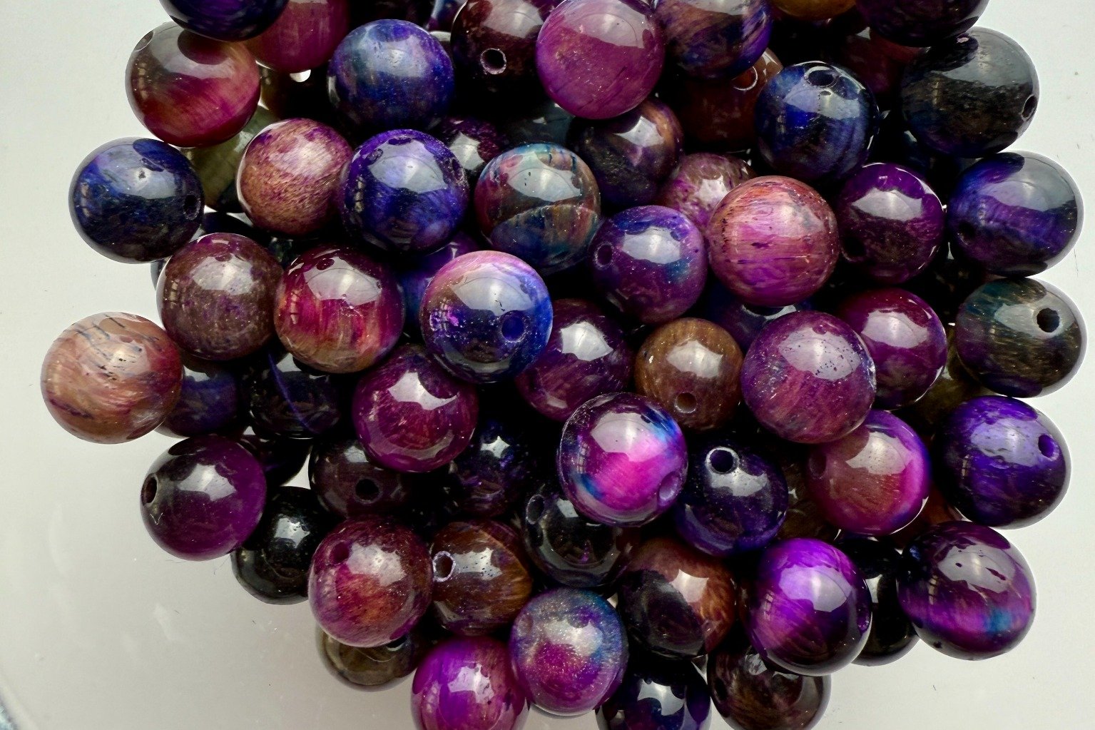Tigerauge-Perlen in faszinierender Galaxy-Mischung aus Violett, Blau, Pink und erdigen Braunnuancen, glänzend arrangiert in einer Glasschale.