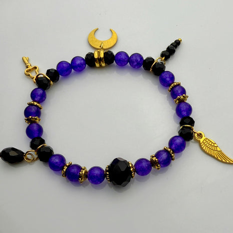 Elegantes Armband ‚Lila Traum‘ mit leuchtend lila Steinperlen, facettierten schwarzen Glasperlen und goldenen Akzenten. Verziert mit Anhängern wie einem goldenen Mond und einem Engelsflügel – ein luxuriöses, handgefertigtes Schmuckstück aus der Schweiz.