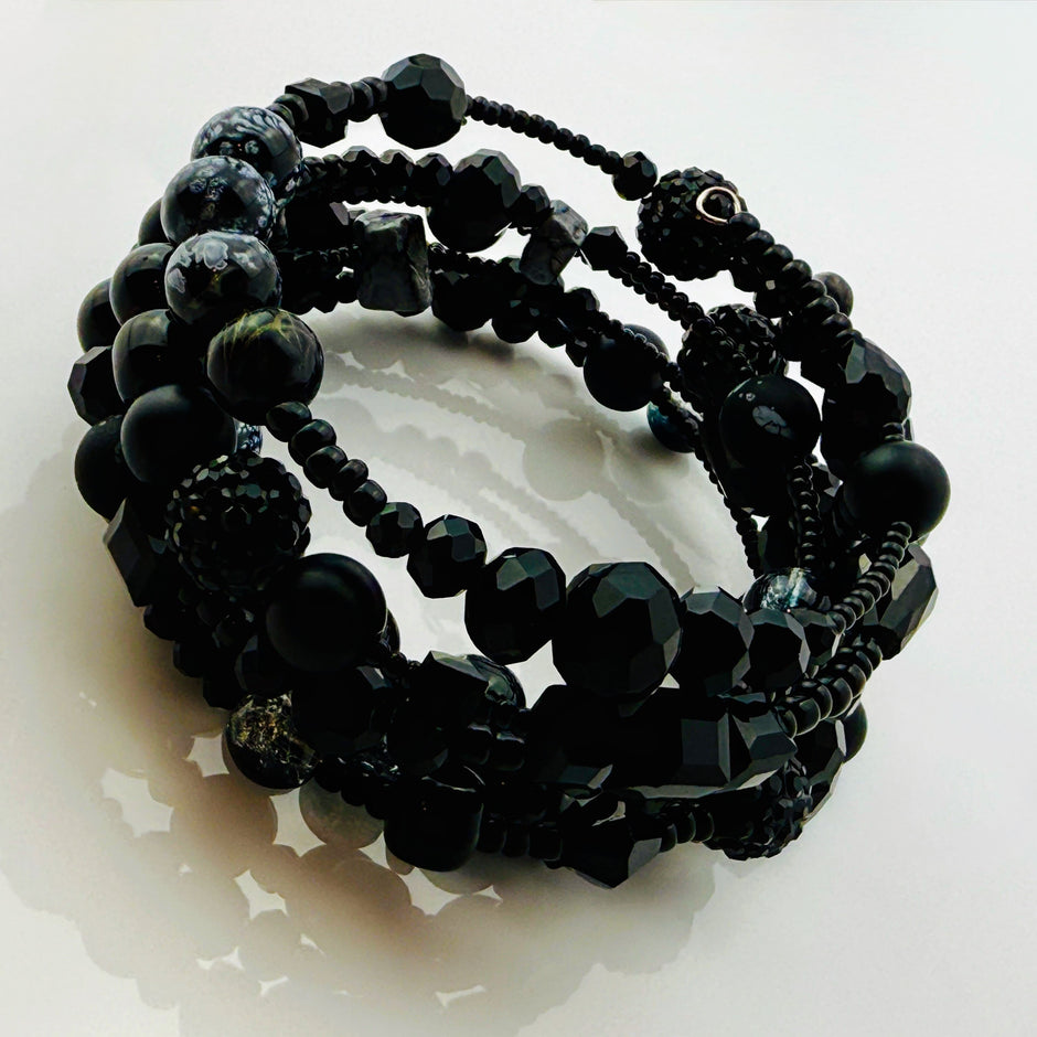 Elegantes spiralförmiges Armband mit einer vielseitigen Kombination aus schwarzen Glasperlen in verschiedenen Größen und Oberflächen. Handgefertigt in der Schweiz, flexibel ohne Verschluss, perfekt für stilvolle Alltagslooks und besondere Anlässe.