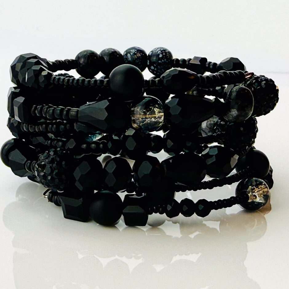 Elegantes spiralförmiges Armband mit einer vielseitigen Kombination aus schwarzen Glasperlen in verschiedenen Größen und Oberflächen. Handgefertigt in der Schweiz, flexibel ohne Verschluss, perfekt für stilvolle Alltagslooks und besondere Anlässe.