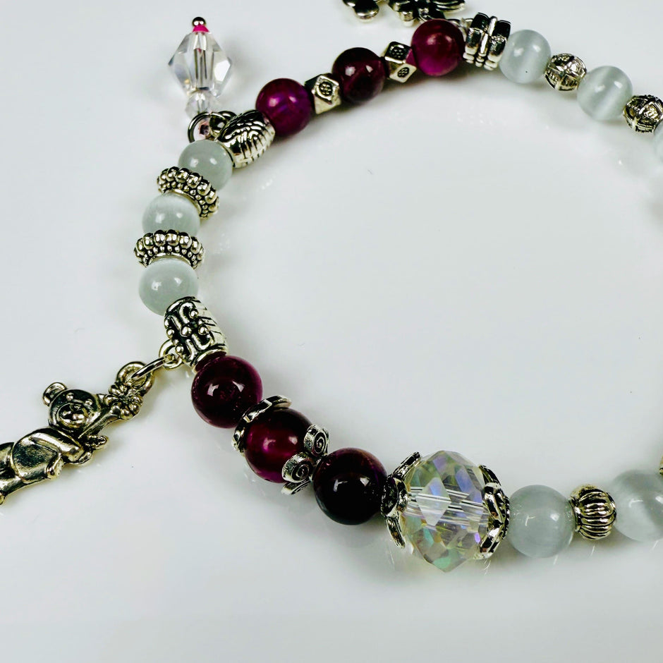 Verspieltes Damenarmband mit Anhängern wie Teddybär, Blüten und Schmuckelementen, kombiniert mit weißen und weinroten Perlen, ideal als verspieltes Accessoire.