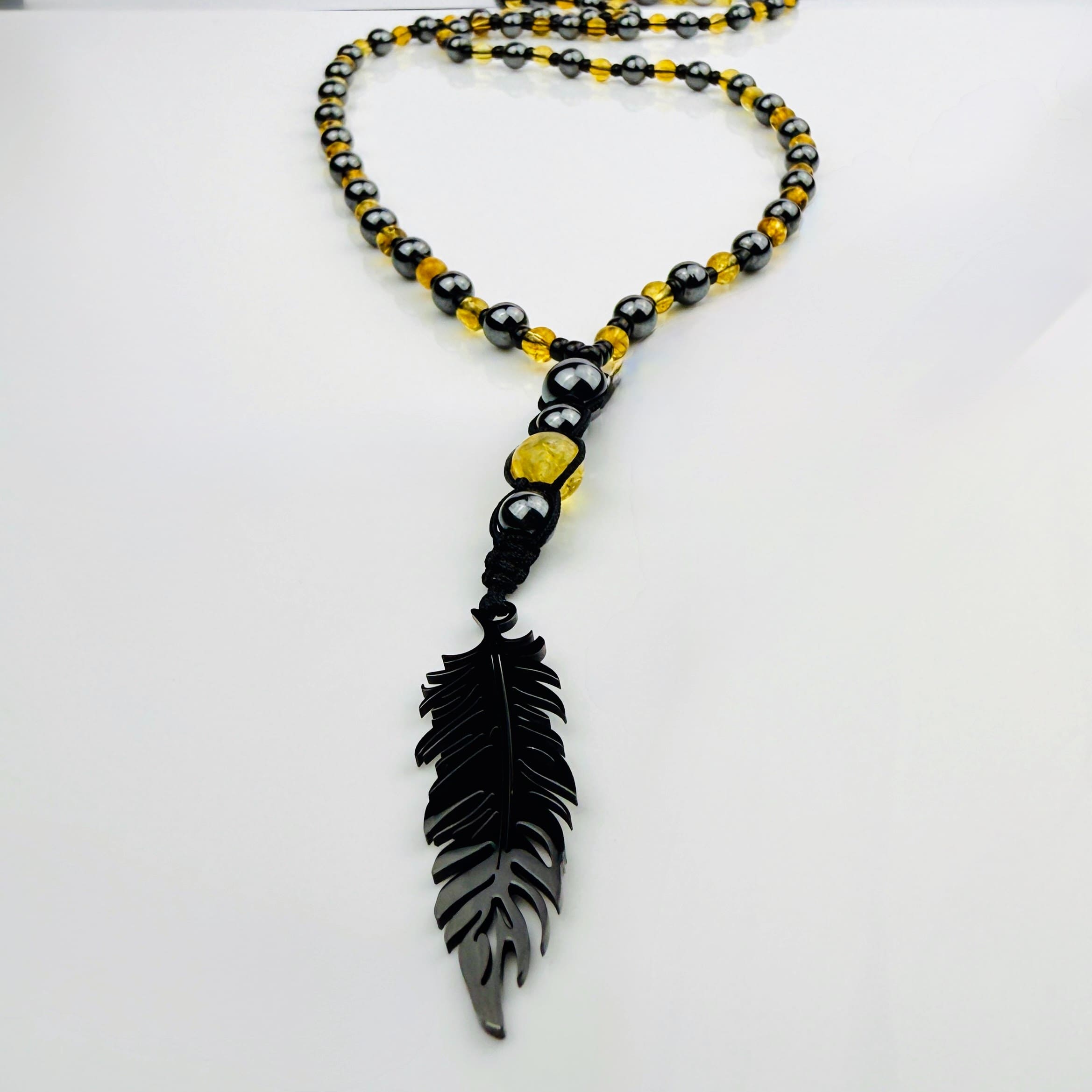 Handgefertigte Y-Halskette für Herren mit gelbem Citrin, Hämatit-Perlen und einem eleganten Edelstahl-Federnanhänger. Kunstvolle Makramee-Technik und schwarze Rocailles ergänzen das moderne und stilvolle Design.