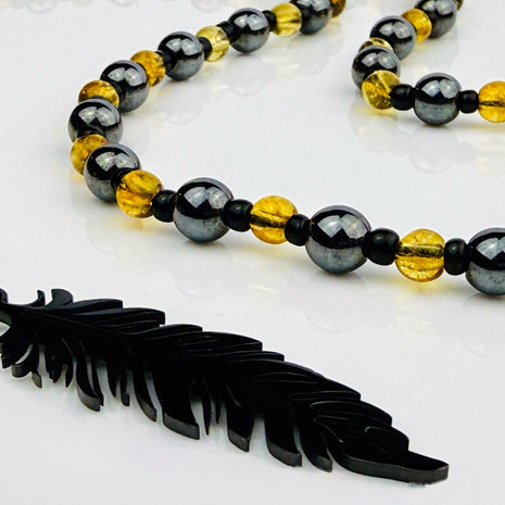 Handgefertigte Y-Halskette für Herren mit gelbem Citrin, Hämatit-Perlen und einem eleganten Edelstahl-Federnanhänger. Kunstvolle Makramee-Technik und schwarze Rocailles ergänzen das moderne und stilvolle Design.